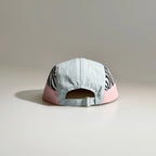 Back view of a light blue and pink five-panel cap with zebra print side panels, an open back and adjustable metal buckle strap.