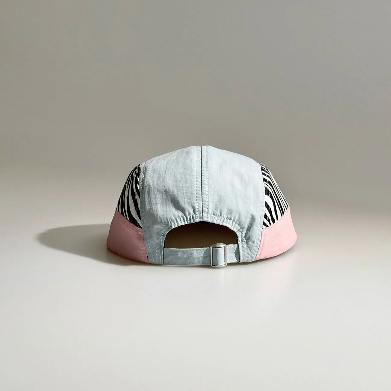 Back view of a light blue and pink five-panel cap with zebra print side panels, an open back and adjustable metal buckle strap.