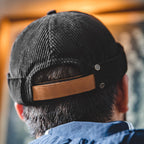 Back view of a black cotton corduroy brimless docker cap with brown adjustable strap and metal studs