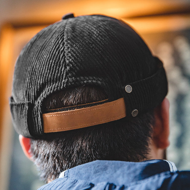Back view of a black cotton corduroy brimless docker cap with brown adjustable strap and metal studs