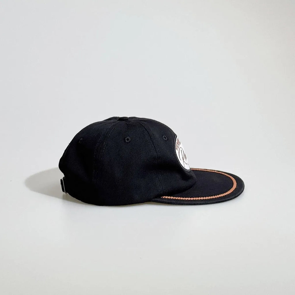 Side view of a black rope cap showing the panelled crown, embroidered racing patch, tan brim trim and adjustable back strap.