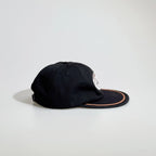Side view of a black rope cap showing the panelled crown, embroidered racing patch, tan brim trim and adjustable back strap.