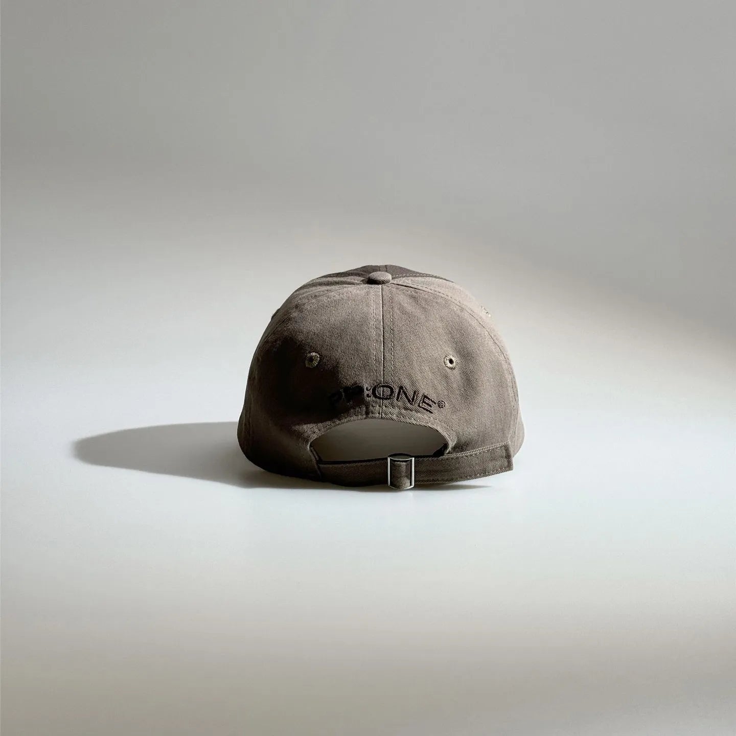 Back view of a brown adjustable baseball cap with embroidered lettering, ventilation eyelets and a metal buckle strap.