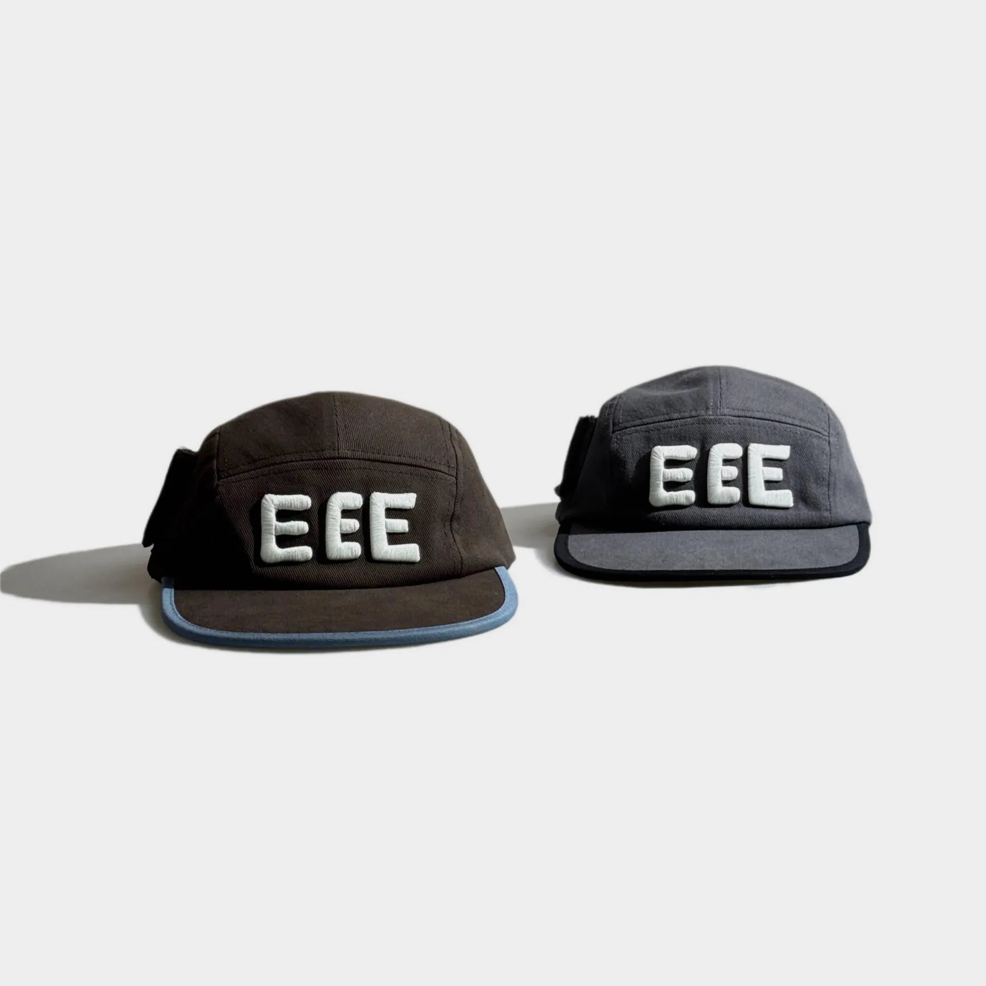 Top view of brown and grey five-panel caps with raised white EEE embroidery, flat brims and contrast edge trim.