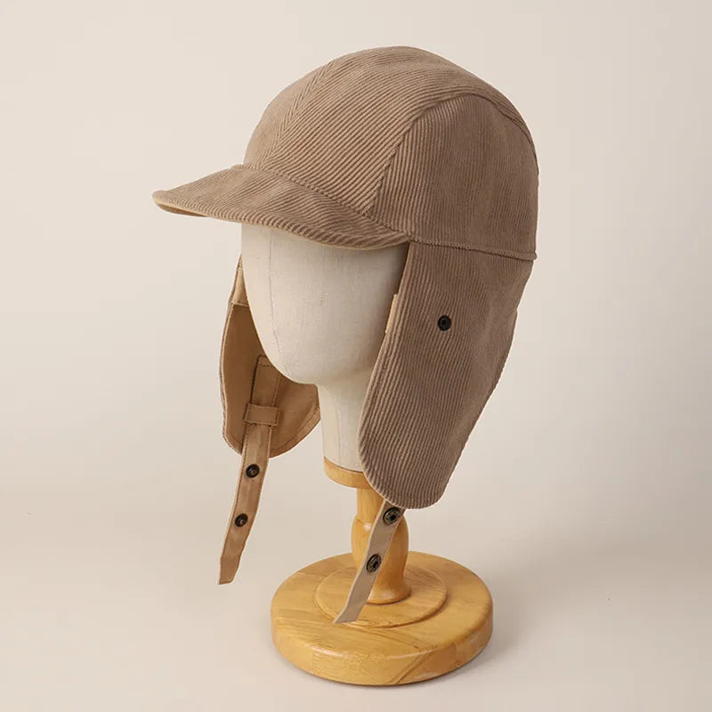 Beige corduroy ear flap cap on a display head with ribbed panels, curved peak and hanging chin straps.