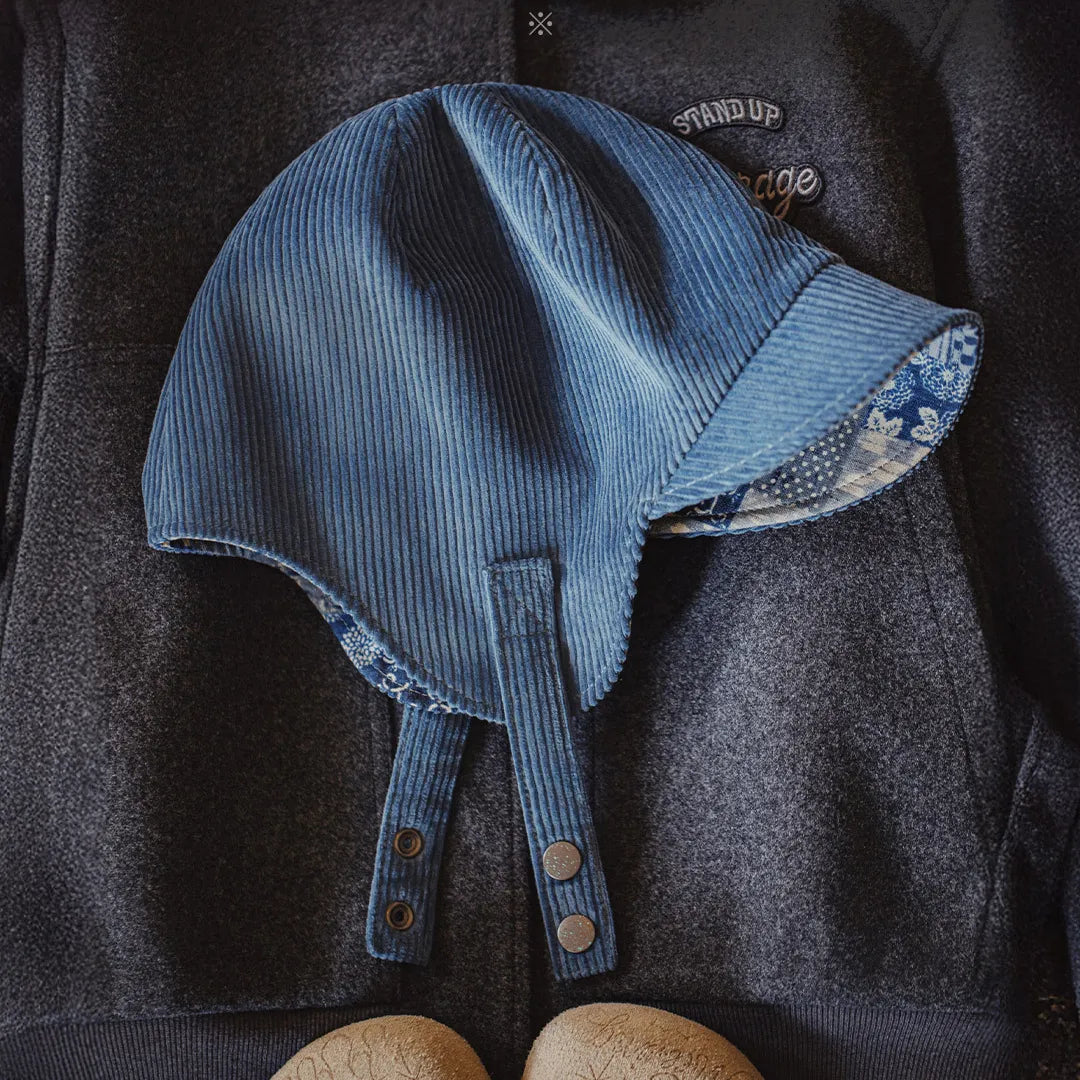 Flat lay of a navy cotton corduroy ear flap cap with patterned under-brim and metal snap straps