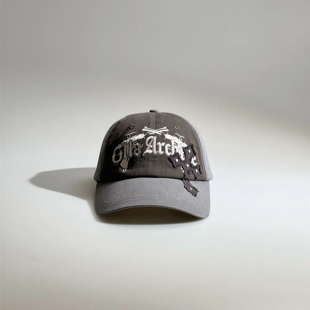 Front view of a grey gothic embroidered baseball cap with layered white lettering, patch details and a curved brim.