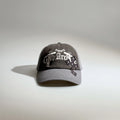 Front view of a grey gothic embroidered baseball cap with layered white lettering, patch details and a curved brim.