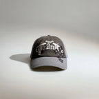 Front view of a grey gothic embroidered baseball cap with layered white lettering, patch details and a curved brim.