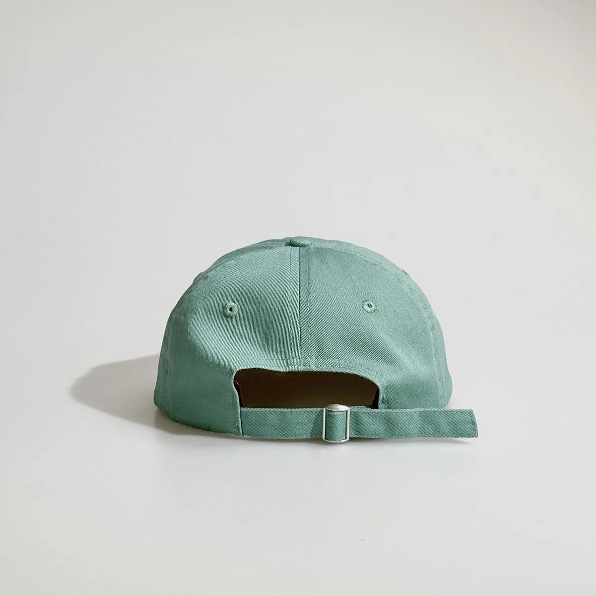 Back view of a mint green baseball cap with embroidered eyelets, an open back and an adjustable metal buckle strap.