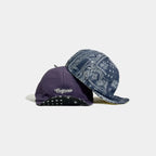 Product shot of two cotton cycling caps: a purple short brim cap with script logo detail and a blue paisley print short brim cap on a clean background.