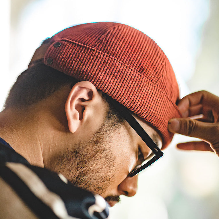 Side view of a man wearing a rust cotton corduroy brimless docker hat with a folded cuff