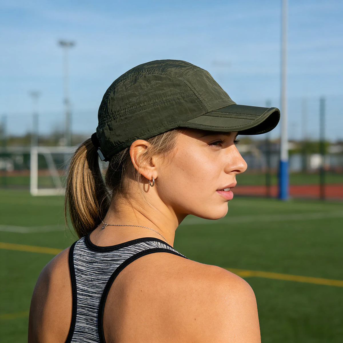 Waterproof baseball cap in olive green, worn by a woman with a ponytail at an outdoor athletics field