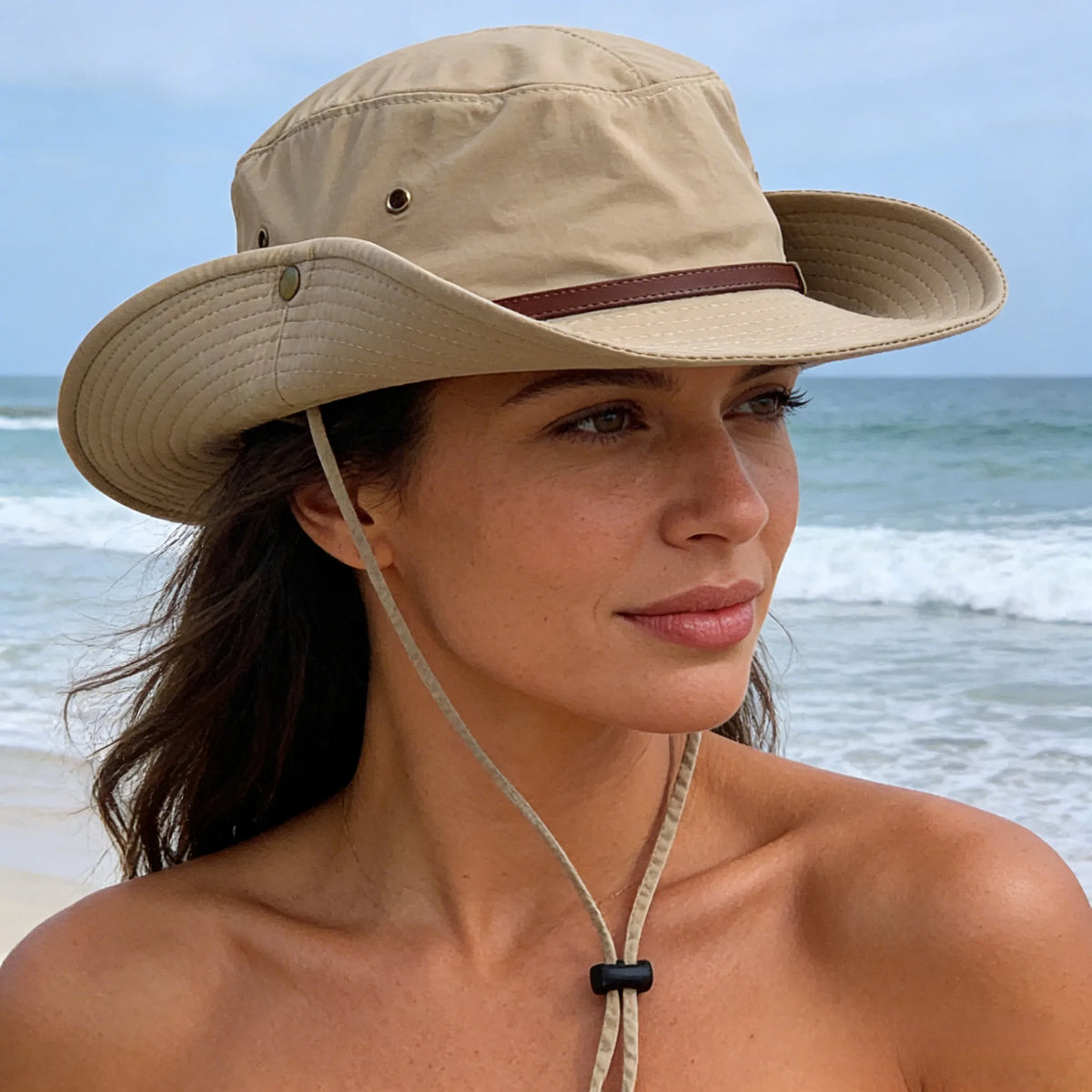 Women's wide brim sun hat in beige worn beside the sea, showing the snap-up side and adjustable chin cord